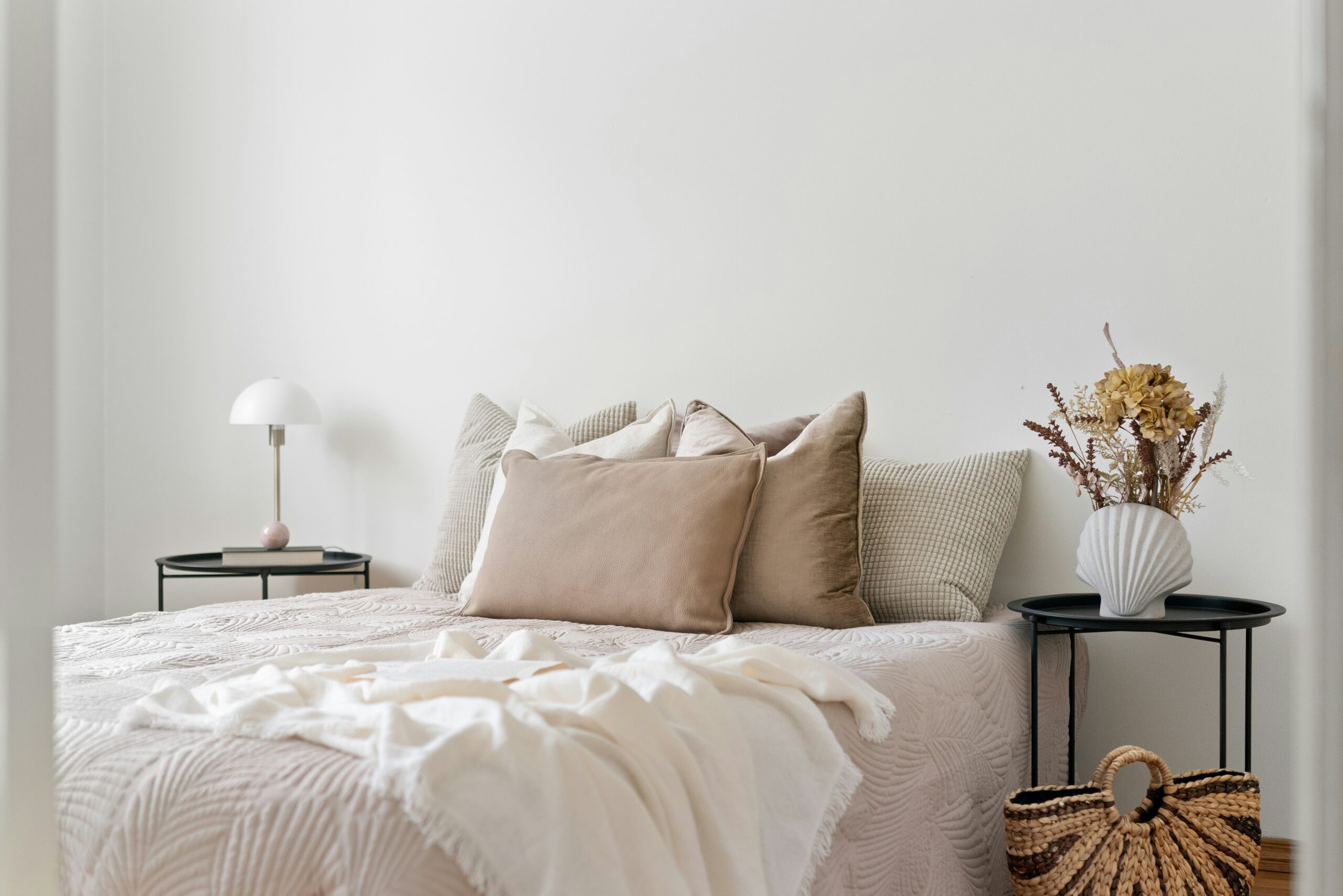 Minimalist bedroom with a neatly made bed featuring neutral-toned pillows, a white throw blanket, a modern black side table with a white lamp, and a decorative vase with dried flowers.